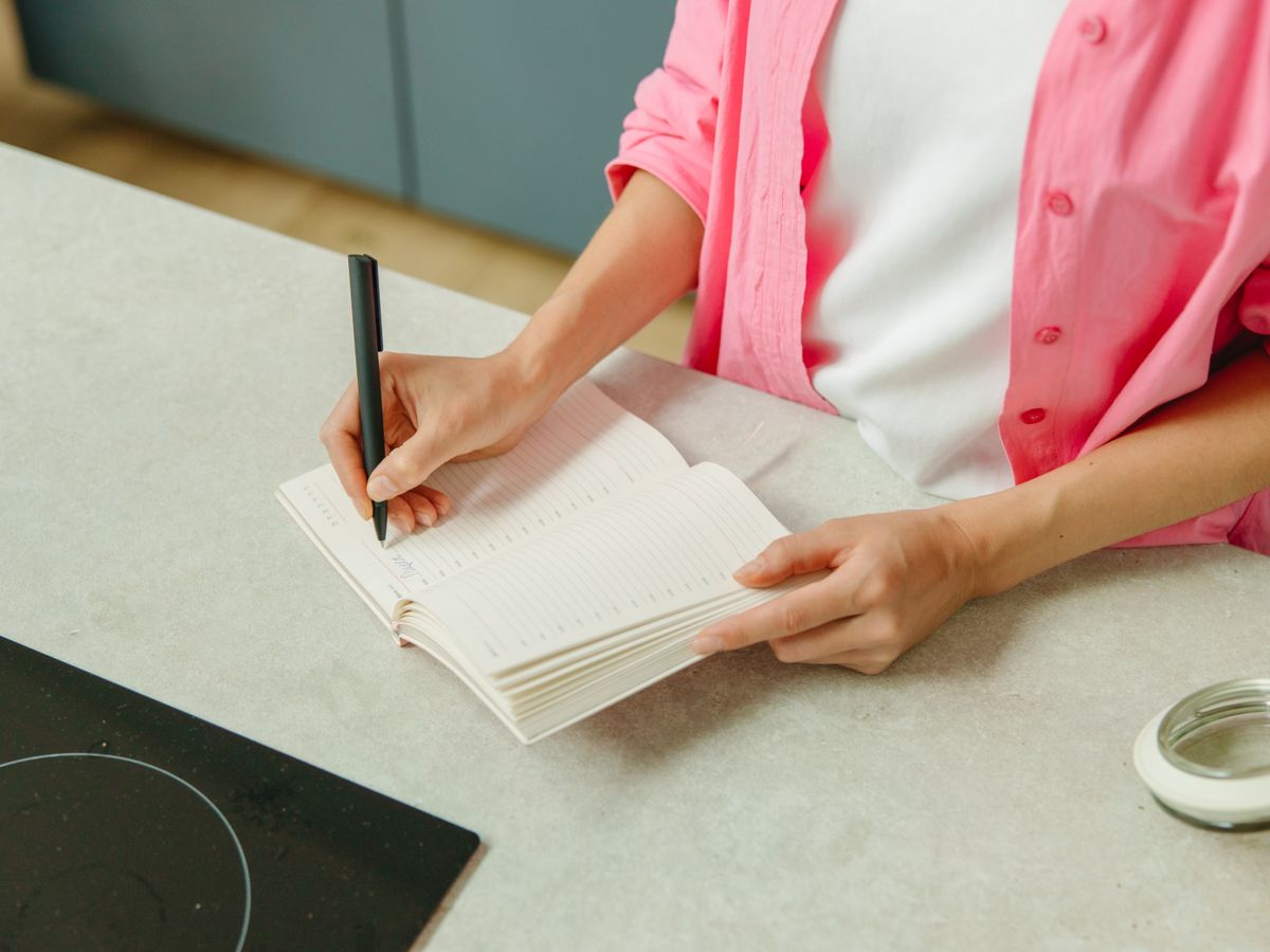 Woman writing a grocery list in a notebook at a kitchen counter before heading to the store